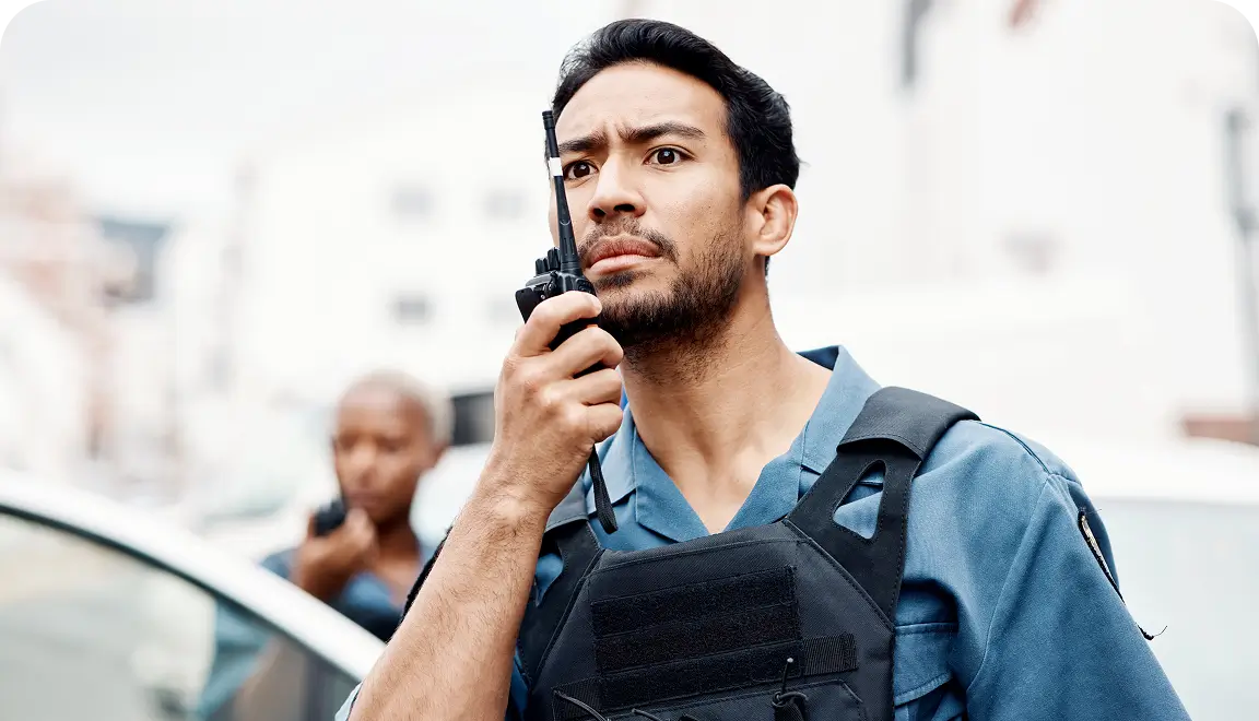 Security officer in a blue uniform with tactical vest speaking into a handheld radio on a city street, focused expression.