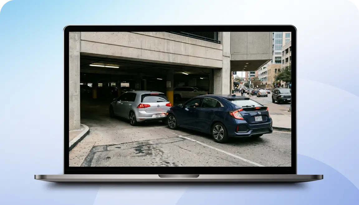 Laptop screen showing a city street with a dark blue sedan reversing from a parking garage, a silver car nearby. (informative)