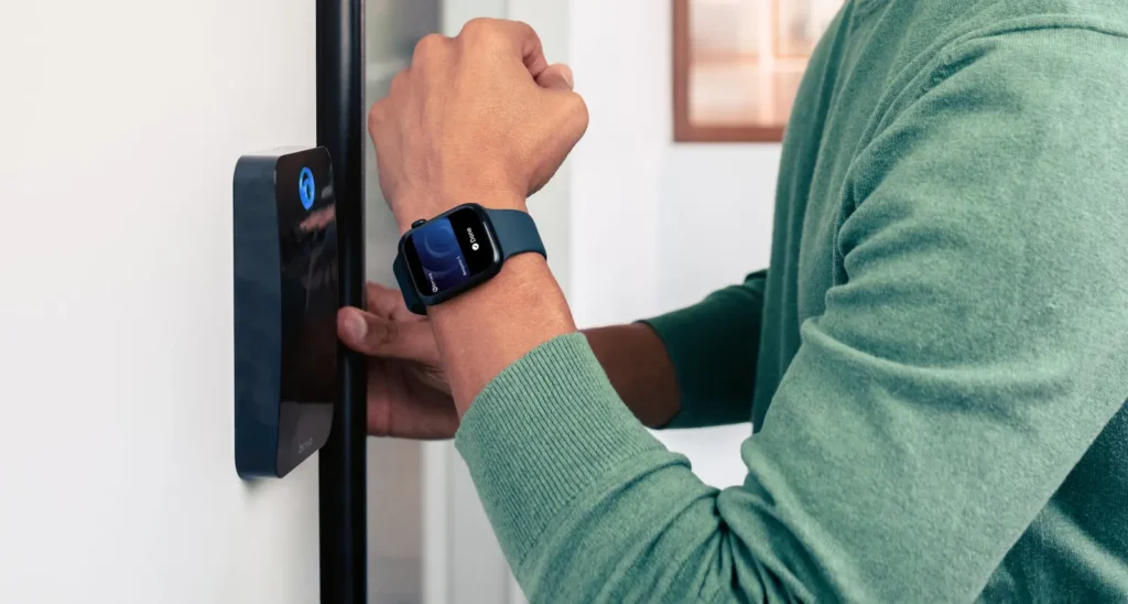 Person in a green sweater wears a blue smartwatch while unlocking a smart door lock on a white interior door.
