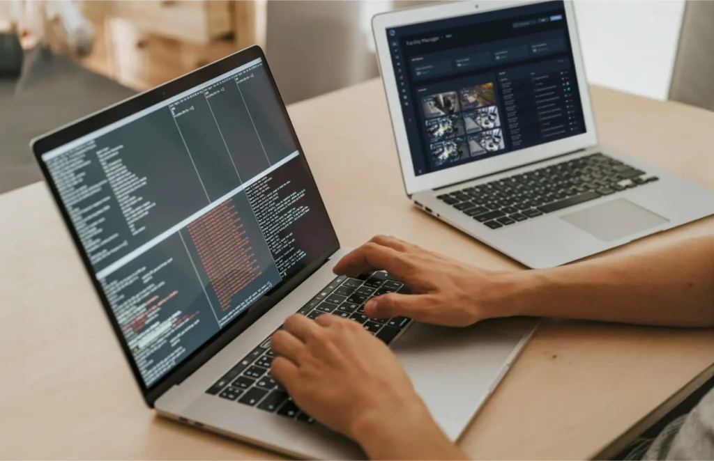 Person typing code on a laptop with a second laptop displaying a dark dashboard in the background.