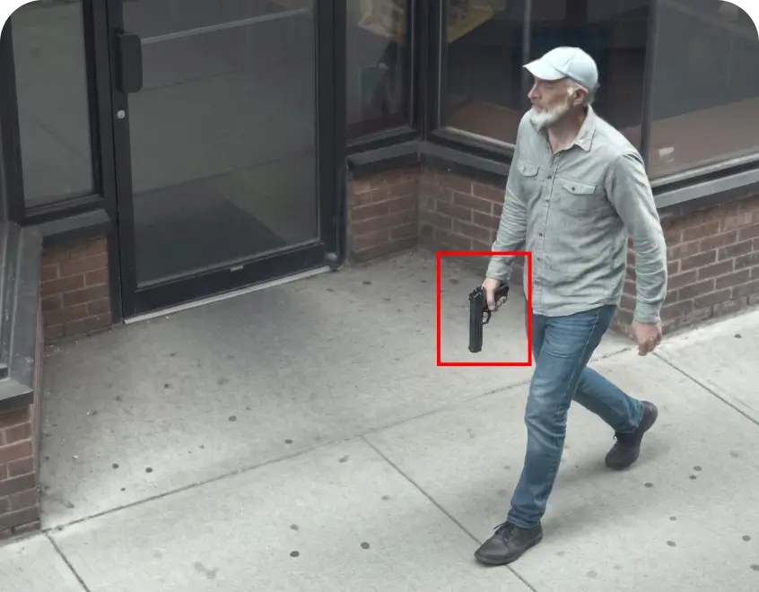 Man in a gray shirt and blue jeans walking on a sidewalk outside a brick storefront, holding a handgun in his right hand.