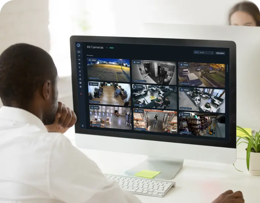 Man seated at a white desk watching a large monitor displaying a grid of security camera feeds in an office environment