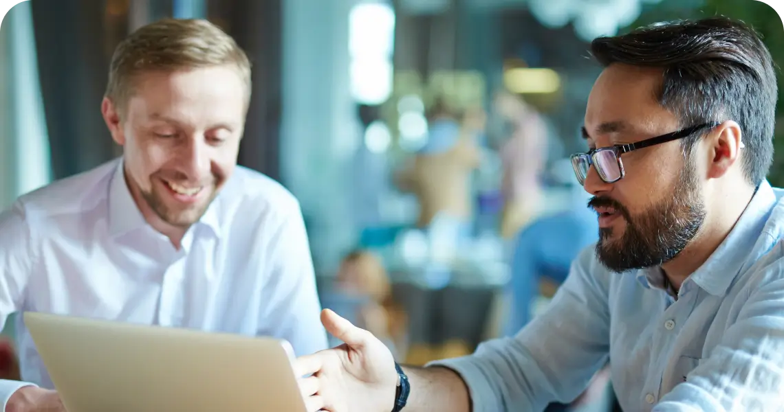 Two men in a bright office sit at a table, smiling as they look at a tablet together (informal meeting).