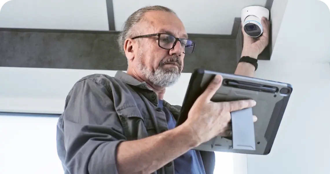 Older man with glasses installing a white security camera on the ceiling while holding a tablet edge-on toward the camera.