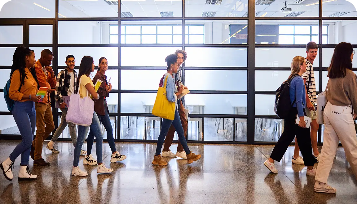Group of students walking through a bright glass-walled campus hallway, carrying backpacks and books.
