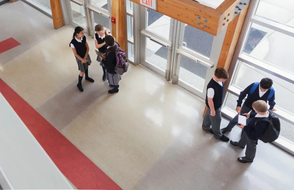 Group of students in uniformes standing in a school hallway near exit doors, talking and looking at a book while a few others use a tablet nearby.