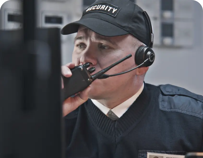 Security guard in a headset speaks into a handheld radio at a monitoring station,