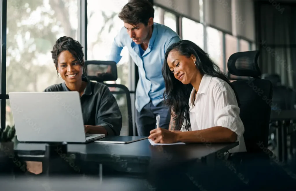 Three colleagues collaborate at a desk: a woman on the left uses a laptop, a woman on the right writes notes, and a man stands behind them looking at the screen.