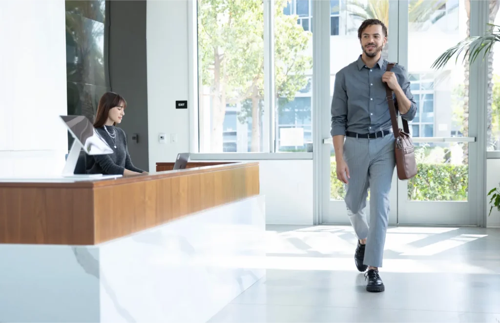 Man in a gray shirt with a brown messenger bag walks into a bright, modern lobby where a receptionist sits at a wooden desk.