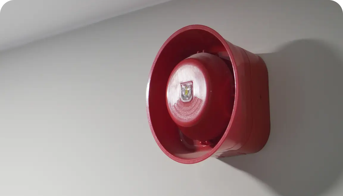 Red, wall-mounted fire alarm bell with a protective circular housing seen from the side (close-up). It signals fire/evacuation alerts.