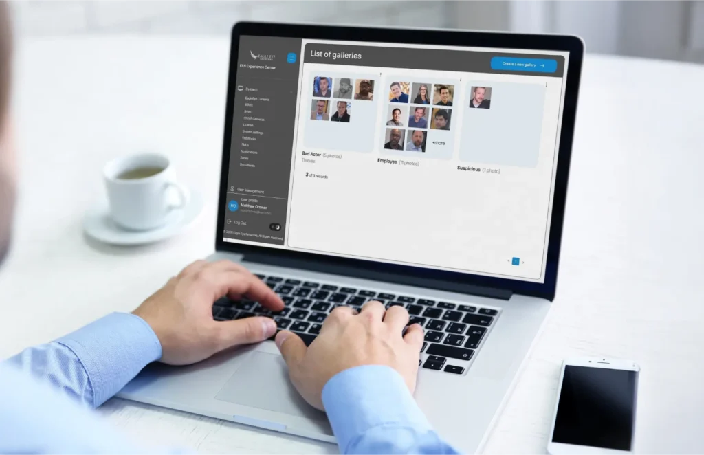 Person typing on a laptop showing a gallery management interface with thumbnail grids on screen, coffee cup nearby