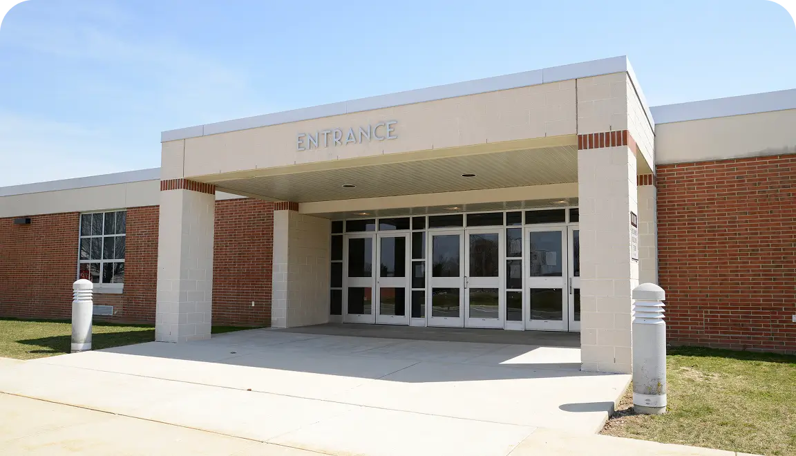 Entrance of a brick-and-beige building with glass doors under a covered drop-off area, labeled 'ENTRANCE'.