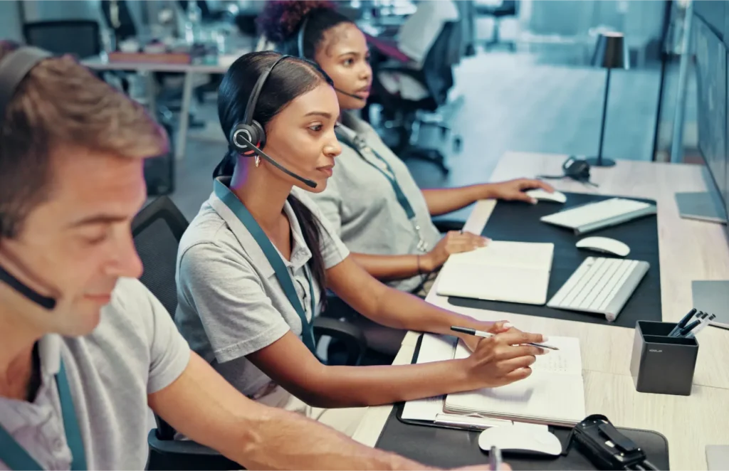 Three call-center agents wear headsets and work at a shared desk with notebooks and keyboards in a bright office.