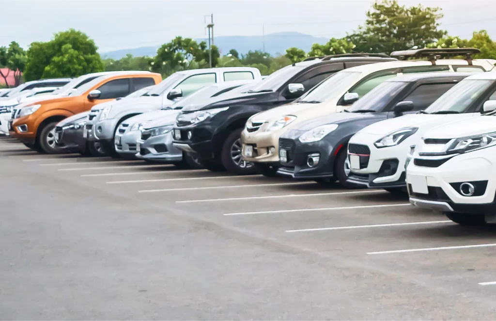 Line of parked cars in a parking lot, various colors with trees in the background.