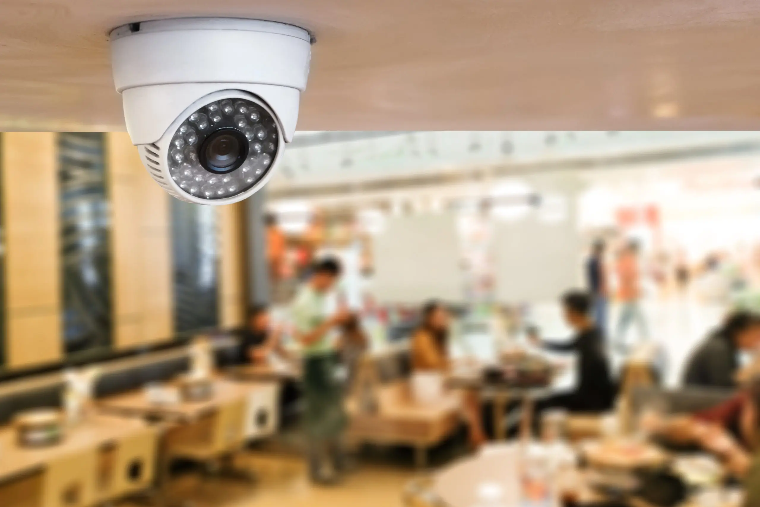 White dome security camera mounted to a ceiling overlooking a busy cafeteria.