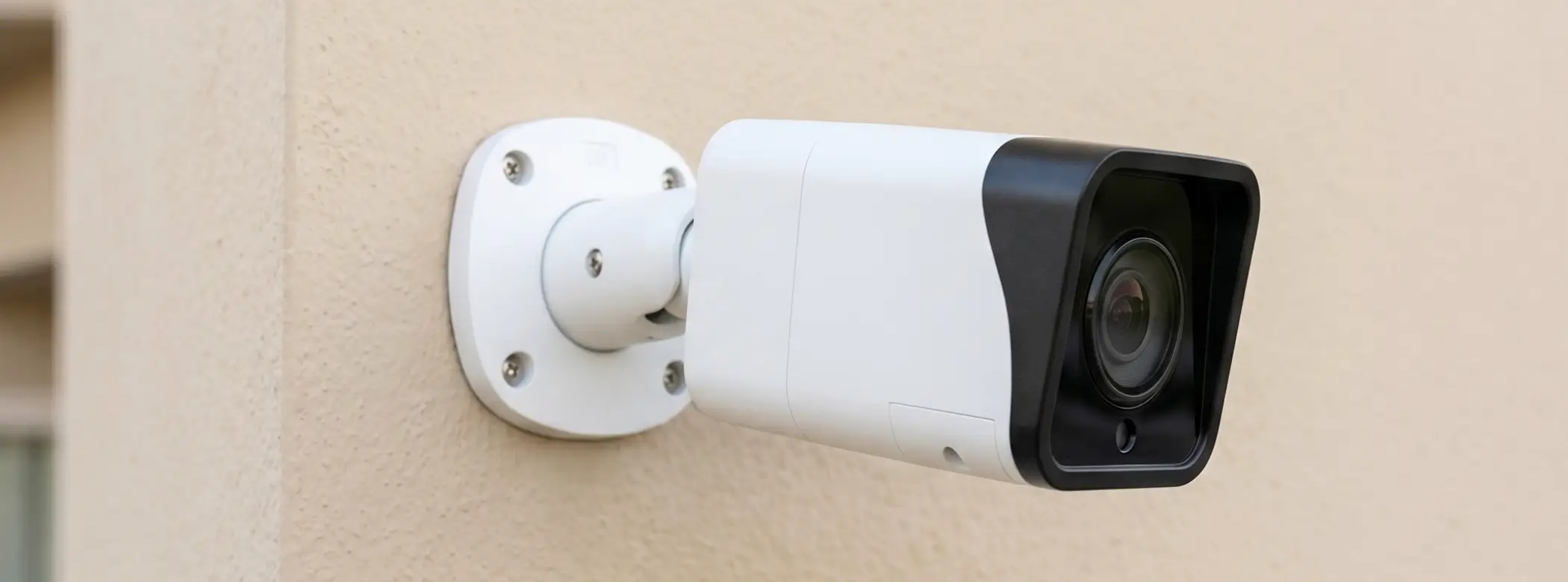 White outdoor surveillance camera mounted on a beige wall, angled to monitor the area