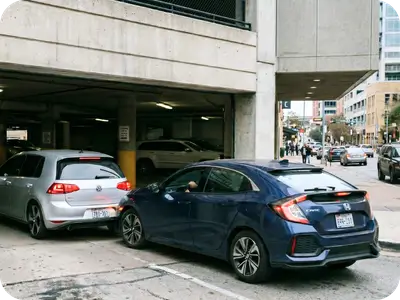 Simply Better Security Blue sedan parked at a curb in front of a concrete parking garage with silver car alongside; city street in background.