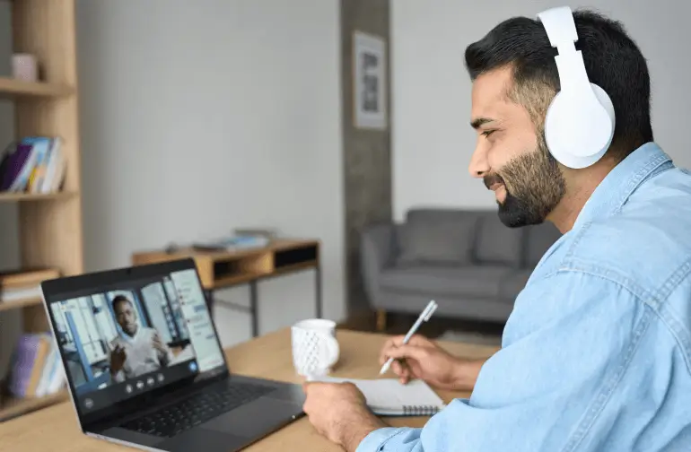 Man wearing white over-ear headphones participates in a video call on his laptop while taking notes in a notebook at a desk with a coffee mug nearby.