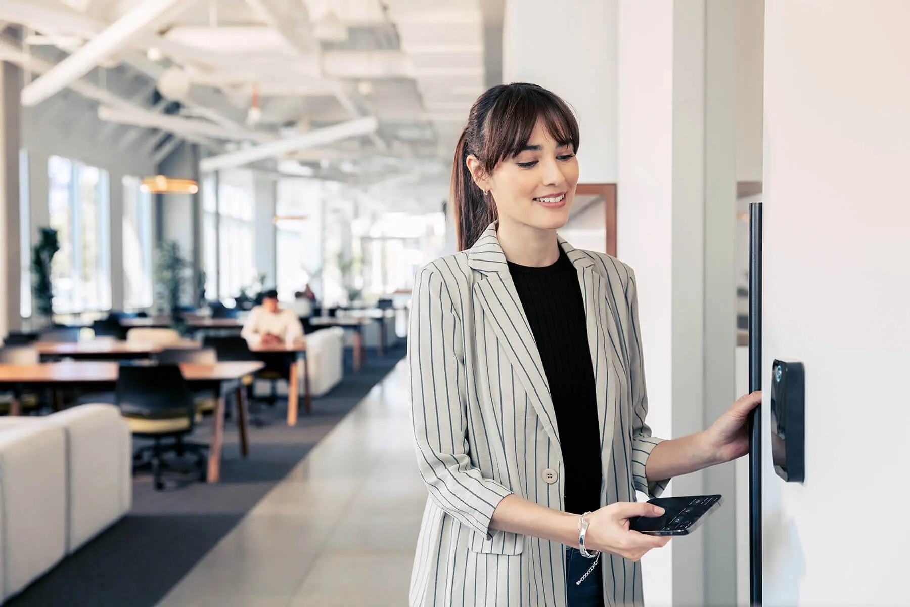 Businesswoman in a striped blazer uses a wall-mounted touchscreen panel in a bright, modern office.