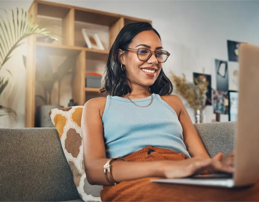 Woman with glasses sitting on a sofa, using a laptop and smiling in a cozy living room setting.