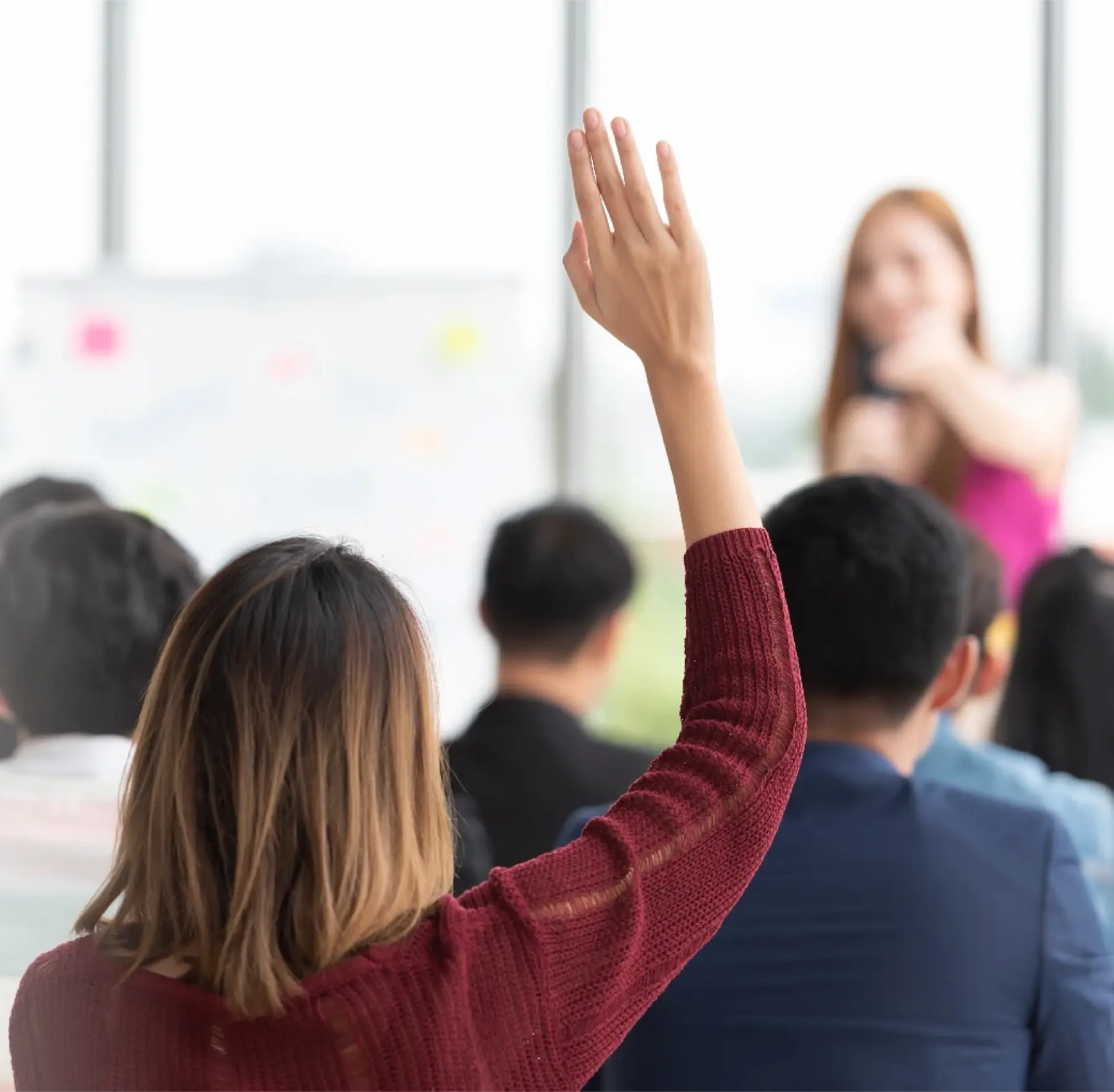 Raised hand of a student in a classroom, indicating participation during a lecture, foreground maroon sweater.