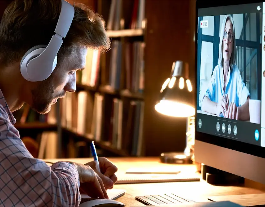 Man wearing white over-ear headphones writes at a desk while a video call shows a woman on the computer screen.