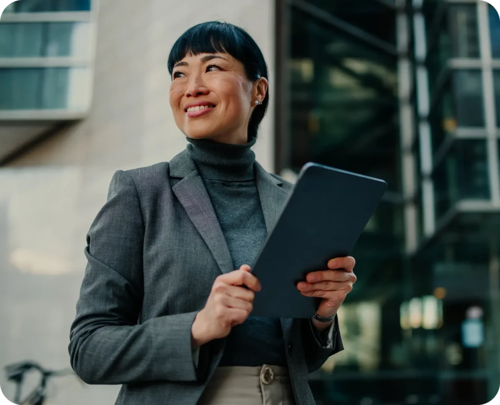 Professional woman in a gray blazer and turtleneck holding a tablet outdoors, smiling and looking upward.